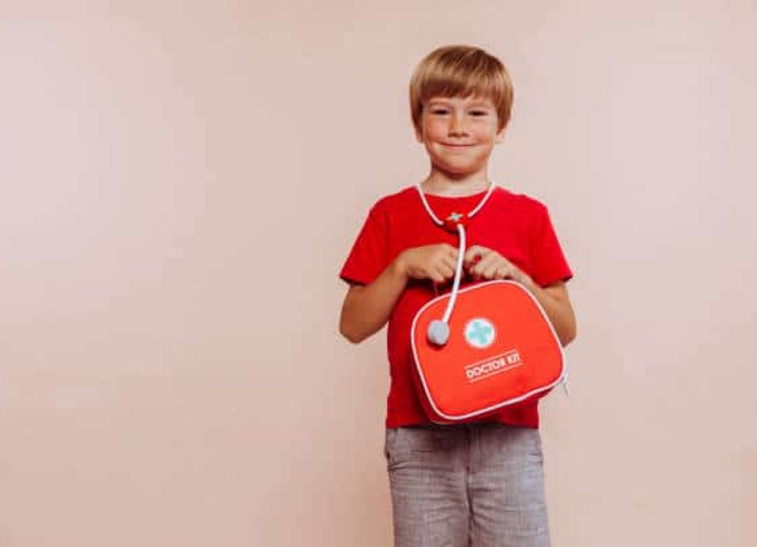 Funny portrait of cute little happy smiling child doctor with first aid kit on hands, wears red t-shirt and studio portrait isolated over beige background. Copy space.